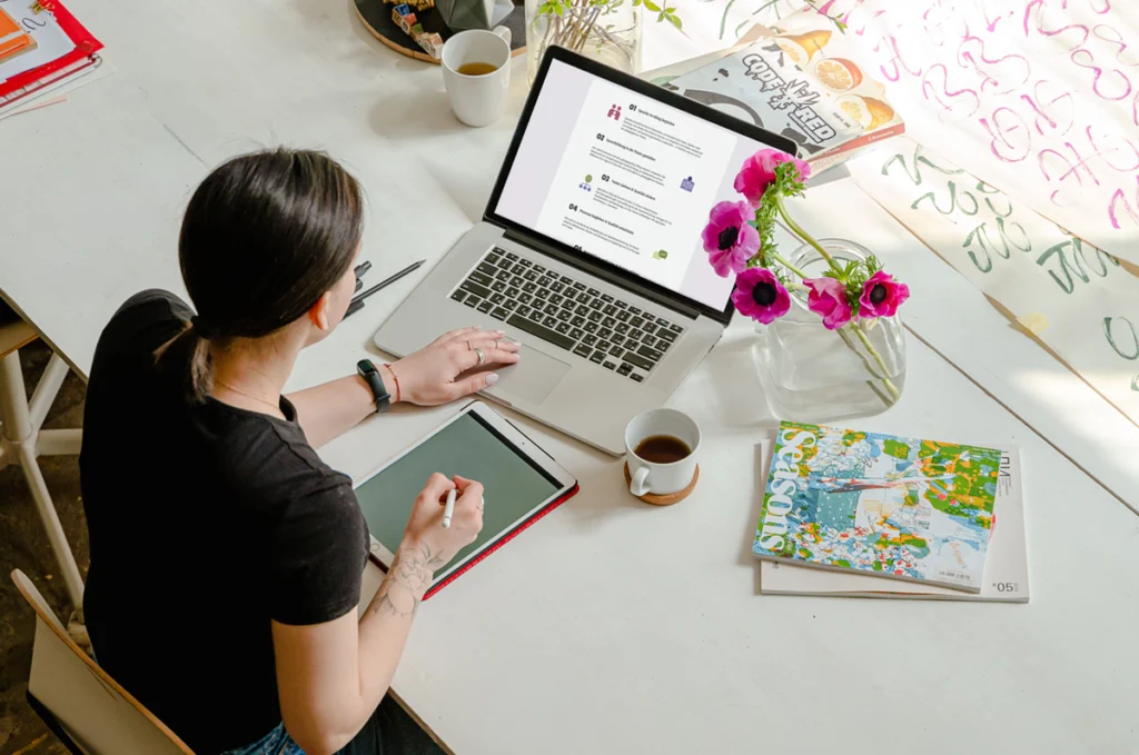 Eine Frau sitzt an einem großen Tisch, auf dem kreative Utensilien verteilt liegen, und arbeitet an einem Laptop auf der Website des Kompetenzzentrums.